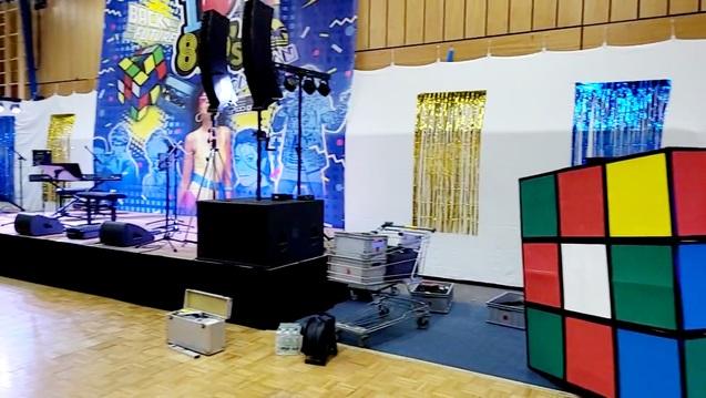 Bühne mit Lautsprechern, einem Klavier und einem großen Rubik's Cube, umgeben von Dekorationen und Ausrüstung.