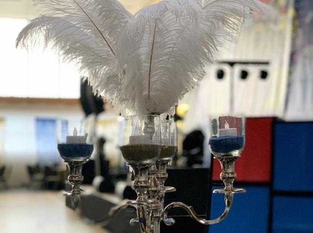 Auf einem silbernen Kerzenständer stehen Gläser mit buntem Sand und Kerzen, überragt von weißen Federn.