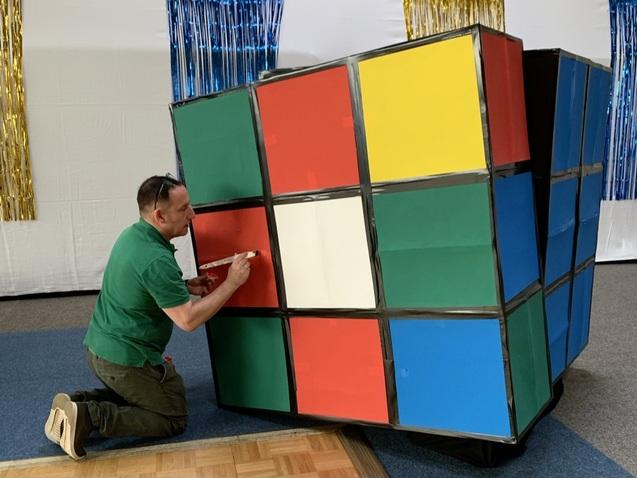 Mann kniet neben einem großen, bunten Würfel und bringt eine Markierung auf der roten Fläche an.
