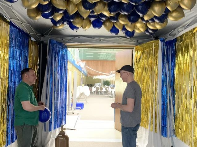 Zwei Männer stehen in einem festlich dekorierten Eingangsbereich mit blauen und goldenen Luftballons.