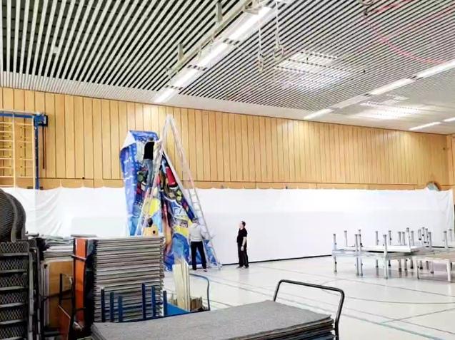 Zwei Personen arbeiten an einer Wand, während eine Leiter mit Dekoration in einer Sporthalle steht. Stapel von Tischen im Vordergrund.