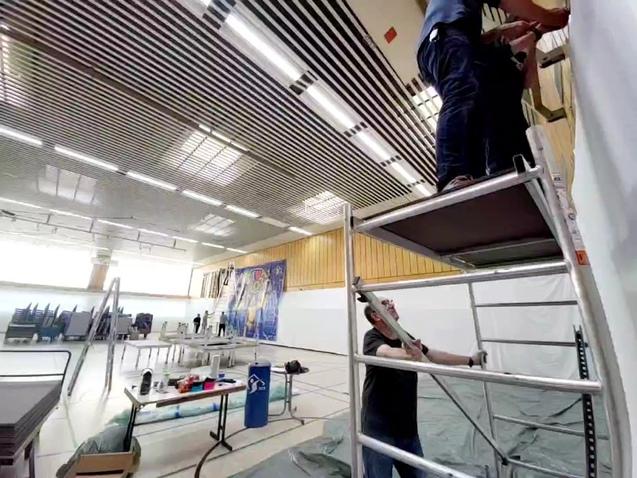 Zwei Personen arbeiten an einer Wand, eine steht auf einer Leiter, während die andere unterstützt. Werkzeug ist sichtbar.