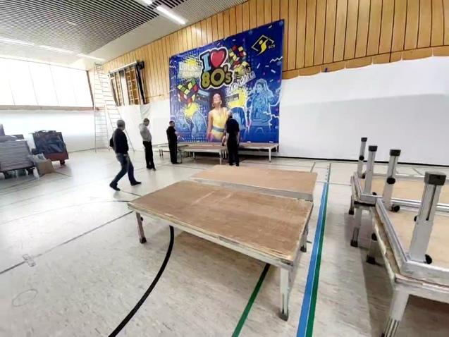 Personen in einer Sporthalle richten eine Bühne mit einem bunten 80er-Jahre-Wandgemälde im Hintergrund auf.