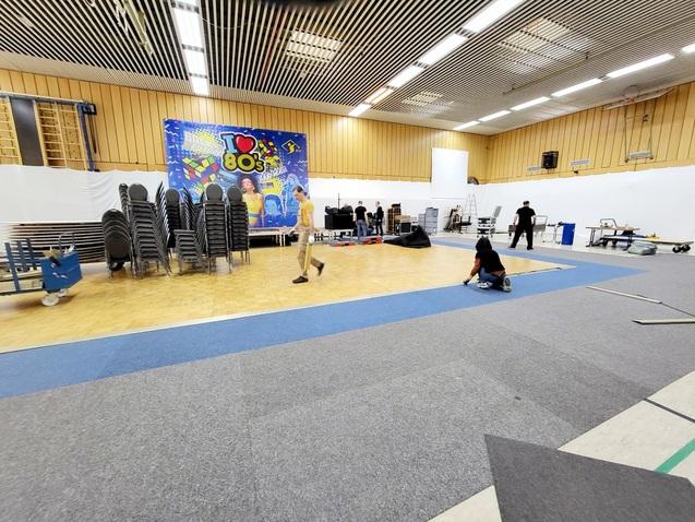 Menschen richten einen Veranstaltungsraum mit Stühlen, Teppich und Bühnenmaterial für eine 80er-Jahre-Party her.