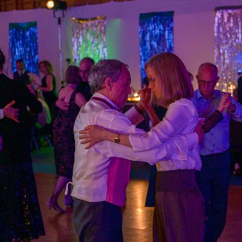 Tanzendes Paar in eleganter Kleidung auf einer beleuchteten Tanzfläche, umgeben von weiteren Feiernden im Hintergrund.