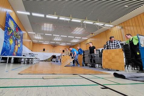 Fünf Personen verlegen Bodenplatten in einer Halle mit buntem Wandbanner im Hintergrund. Tische und Stühle sind aufgestellt.