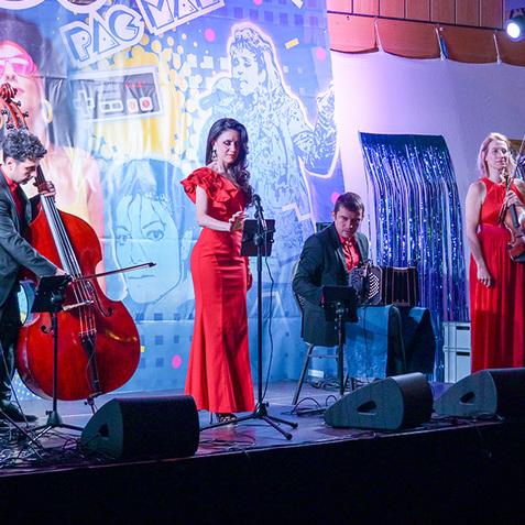 Auftritt einer Musikgruppe in roten Outfits vor einer bunten 80er-Jahre Kulisse mit Mikrofon und Instrumenten.