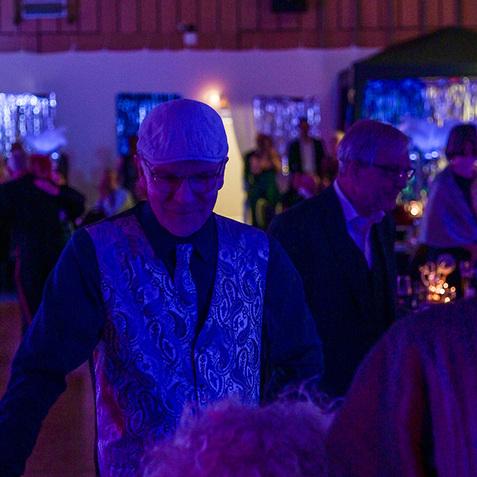 Mann in einem blauen Anzug mit gemusterter Weste steht an einem Tisch in einer festlichen Umgebung mit bunten Lichtern.