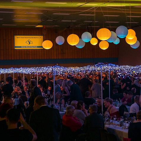 Festliche Veranstaltung in einer Halle mit dekorativen Laternen und Lichtern, Besucher sitzen an Tischen und feiern.