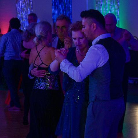 Tanzende Paare in eleganter Kleidung bewegen sich im gedimmten Licht einer festlichen Tanzveranstaltung.