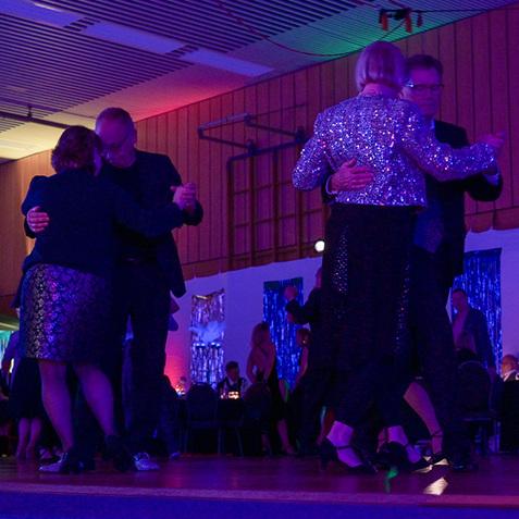 Tanzende Paare in eleganter Kleidung auf einer festlich beleuchteten Tanzfläche in einem Veranstaltungsraum.