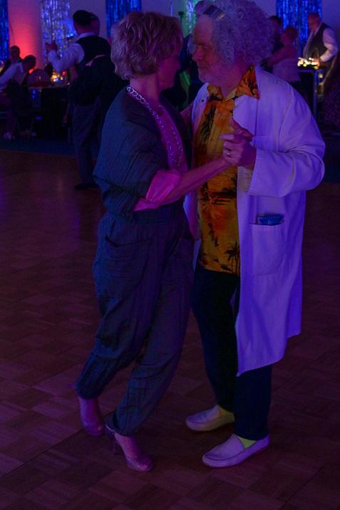 Tanzendes Paar in bunter Beleuchtung, Frau in grauem Anzug und Mann in bunter Hemdbluse mit Laborkittel.