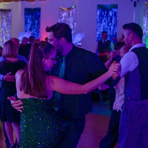 Tanzendes Paar in eleganter Kleidung, umgeben von tanzenden Gästen und bunten Lichtern in einem festlichen Saal.