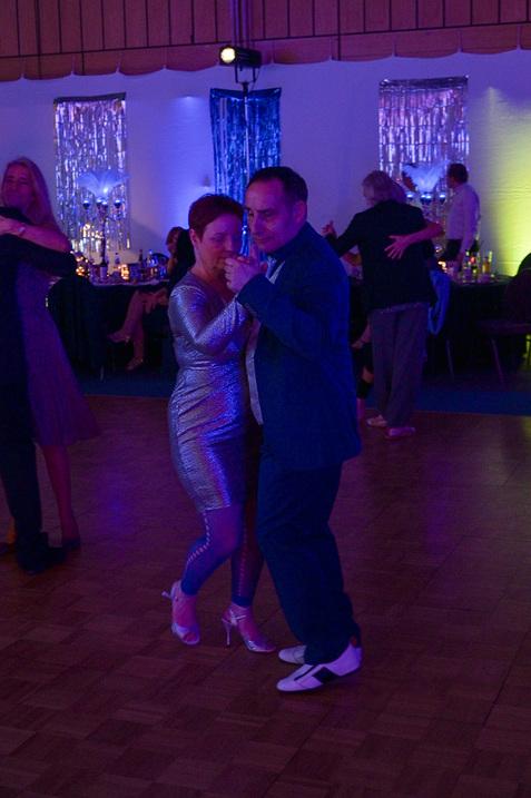 Tanzendes Paar in eleganter Kleidung auf einer beleuchteten Tanzfläche mit anderen Gästen im Hintergrund.