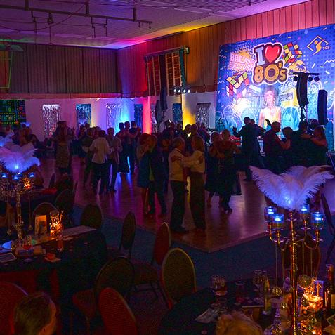 Bunt beleuchteter Saal mit Menschen beim Tanzen, dekoriert im 80er-Jahre-Stil und mit festlichen Tischsets.