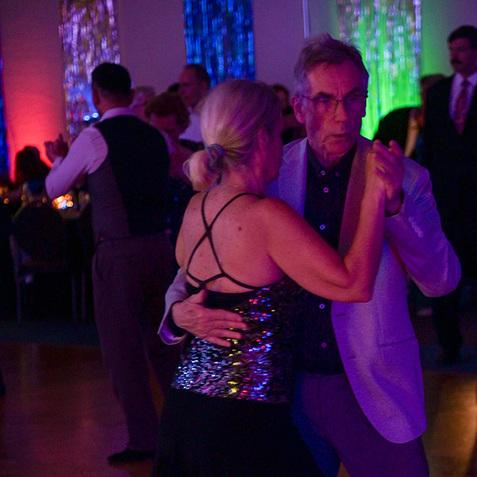 Tanzendes Paar in eleganter Kleidung vor bunten Lichtern auf einer Tanzfläche. Der Mann führt die Frau.