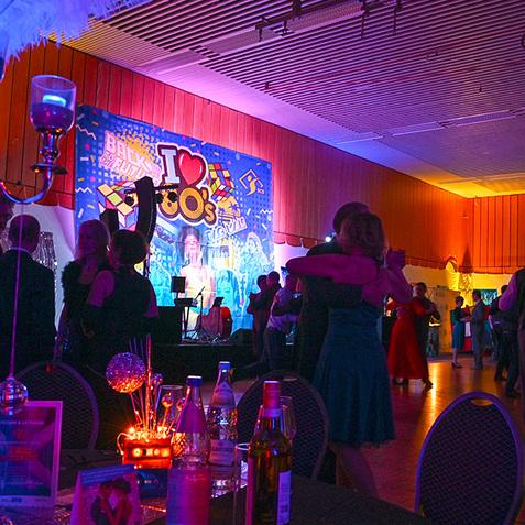 Beleuchteter Veranstaltungsraum mit tanzenden Gästen und festlicher Dekoration im Stil der 80er Jahre.