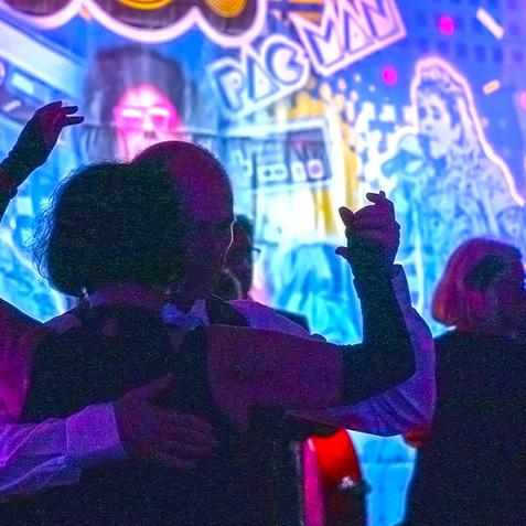 Tanzende Paare vor einer bunten Wanddekoration im 80er-Jahre-Stil mit Motiven von Pac-Man und Retro-Elementen.