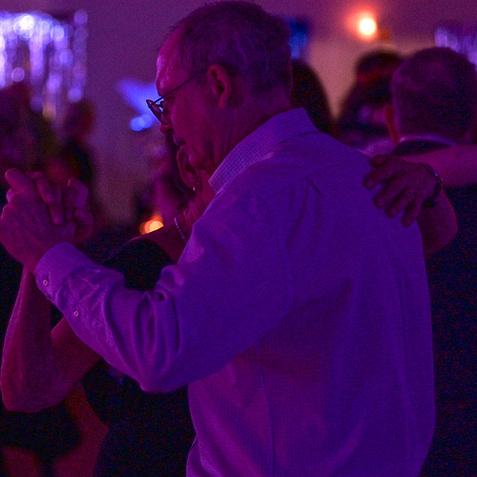 Tanzpaar in einem schwach beleuchteten Saal, umgeben von anderen tanzenden Gästen und bunten Lichtern.