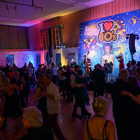 Tanzende Gäste auf einer 80er-Jahre-Party mit bunten Lichtern und einer Band im Hintergrund.