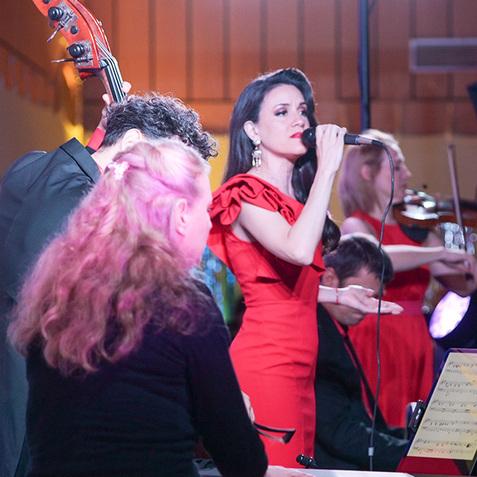 Musiker in roten Outfits stehen auf einer Bühne, während eine Sängerin mit Mikrofon ein Lied vorträgt.