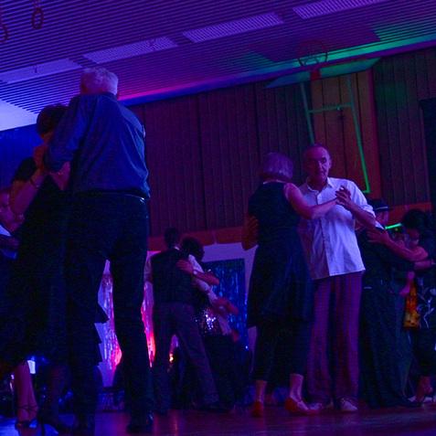 Tanzende Paare in eleganter Kleidung in einem farbig beleuchteten Raum während einer Tanzveranstaltung.