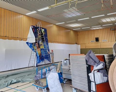 Drei Personen hängen ein großes Banner an eine Wand in einer Sporthalle, umgeben von aufgerollten Teppichen und Gerätschaften.