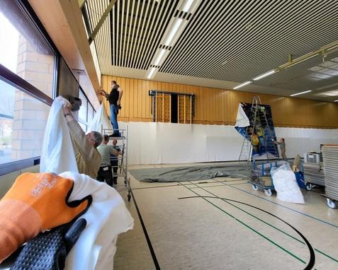 Arbeiter bringen weiße Abdeckungen in einer Sporthalle an, während Leitern und Materialien im Hintergrund stehen.