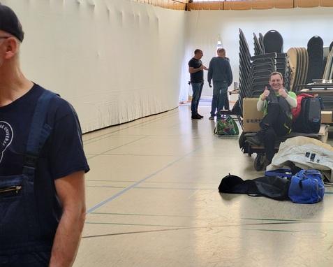 Personen in einer Sporthalle, ein Mann posiert mit Daumen hoch, Stühle und Taschen im Hintergrund.