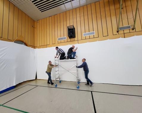 Vier Personen montieren ein weißes Tuch mit einer Leiter in einer Sporthalle, die Wände sind hellbraun vertäfelt.