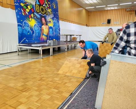 Menschen verlegen Holzpaneele auf einem Tanzboden in einer Halle mit buntem 80er-Jahre-Motiv im Hintergrund.