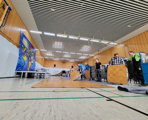 Fünf Personen verlegen Bodenplatten in einer Halle mit buntem Wandbanner im Hintergrund. Tische und Stühle sind aufgestellt.