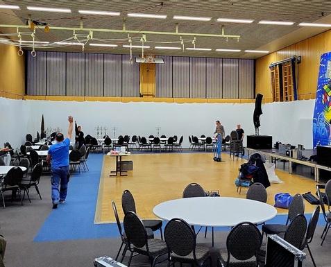 Raum mit runden Tischen und Stühlen, zwei Personen im Hintergrund arbeiten an der Dekoration einer Veranstaltung.