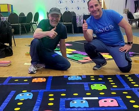 Zwei Männer zeigen Daumen hoch neben einem bunten Pac-Man-Spielbrett auf dem Boden.