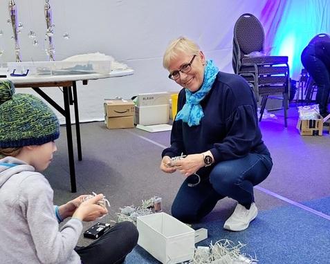 Frau mit Brille und buntem Schal kniet lächelnd neben einem Kind, das mit Bastelmaterial arbeitet, in einem festlich dekorierten Raum.