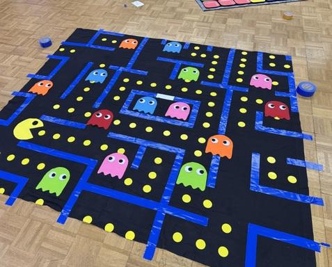 Bunte Spielmatte im Pac-Man-Stil mit Geistern und gelben Punkten auf schwarzem Hintergrund.