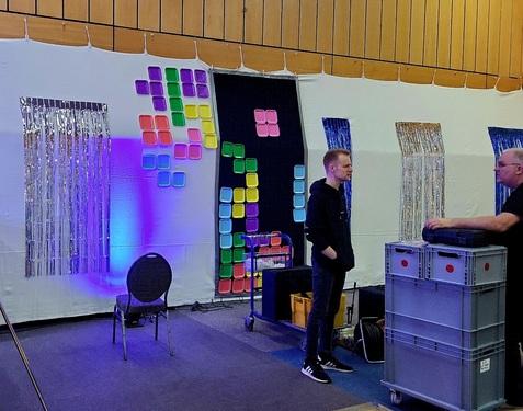 Zwei Personen stehen in einem Raum mit bunten Wanddekorationen und einem Stuhl, während Kisten um sie herum stehen.