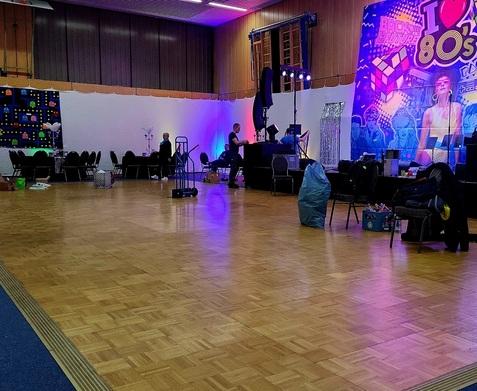 Veranstaltungsraum mit Tanzfläche, bunten Lichtern, Stühlen und einem dekorierten Hintergrund mit 80er-Jahre-Motiven.