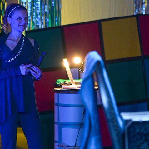 Frau mit Playing-Boards in schimmerndem Licht, trägt ein langes, dunkelblaues Outfit, steht neben einem Tisch.