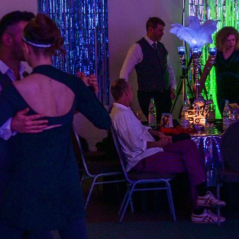 Tanzendes Paar auf einer festlichen Veranstaltung, umgeben von bunten Lichtern und Gästen an Tischen.