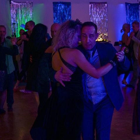 Tanzpaar in eleganter Kleidung tanzt in einem beleuchteten Raum mit anderen Gästen im Hintergrund.