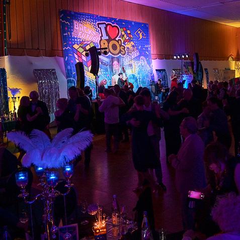 Feierliche Tanzveranstaltung mit festlich gekleideten Gästen und einer bunten 90er-Jahre-Dekoration im Hintergrund.