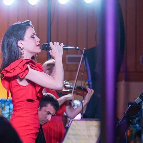 Sängerin in rotem Kleid hält Mikrofon, umgeben von Musikern auf der Bühne mit bunten Lichtern im Hintergrund.