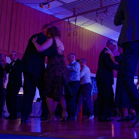 Tanzende Paare in einem festlich beleuchteten Saal, umgeben von Holzvertäfelung und bunten Lichtern.