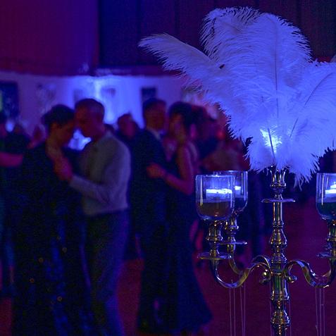 Tisch mit einer eleganten Kerzenhalterung und Federdekoration, im Hintergrund tanzen Menschen in einem beleuchteten Saal.