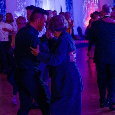 Tanzende Paare in eleganter Kleidung auf einer festlich beleuchteten Tanzfläche mit Holzparkett.