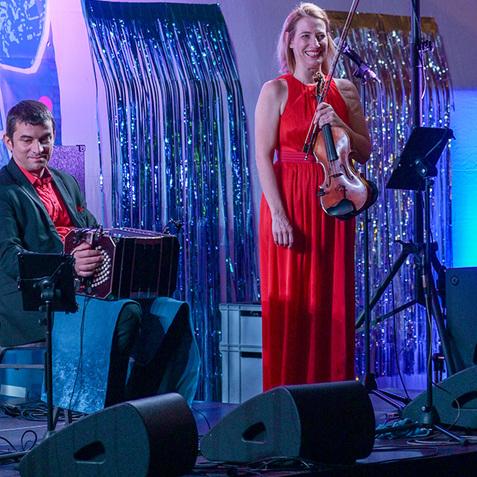 Frau in rotem Kleid hält eine Geige, während ein Mann auf dem Akkordeon spielt; bunte Lichter und Dekoration im Hintergrund.