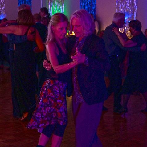 Tanzende Paare in festlicher Kleidung vor einer farbenfrohen, beleuchteten Tanzfläche bei einem Ereignis.