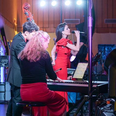 Drei Musiker treten auf einer Bühne auf: Sängerin in rotem Kleid, Pianistin mit roten Rock und Kontrabassist im Hintergrund.