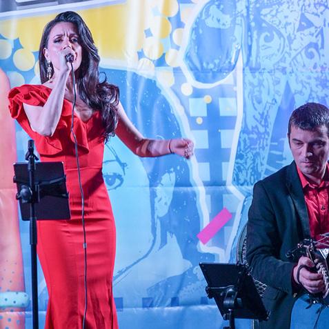 Frau in rotem Kleid singt auf einer Bühne, während ein Musiker neben ihr spielt, vor buntem Hintergrund.